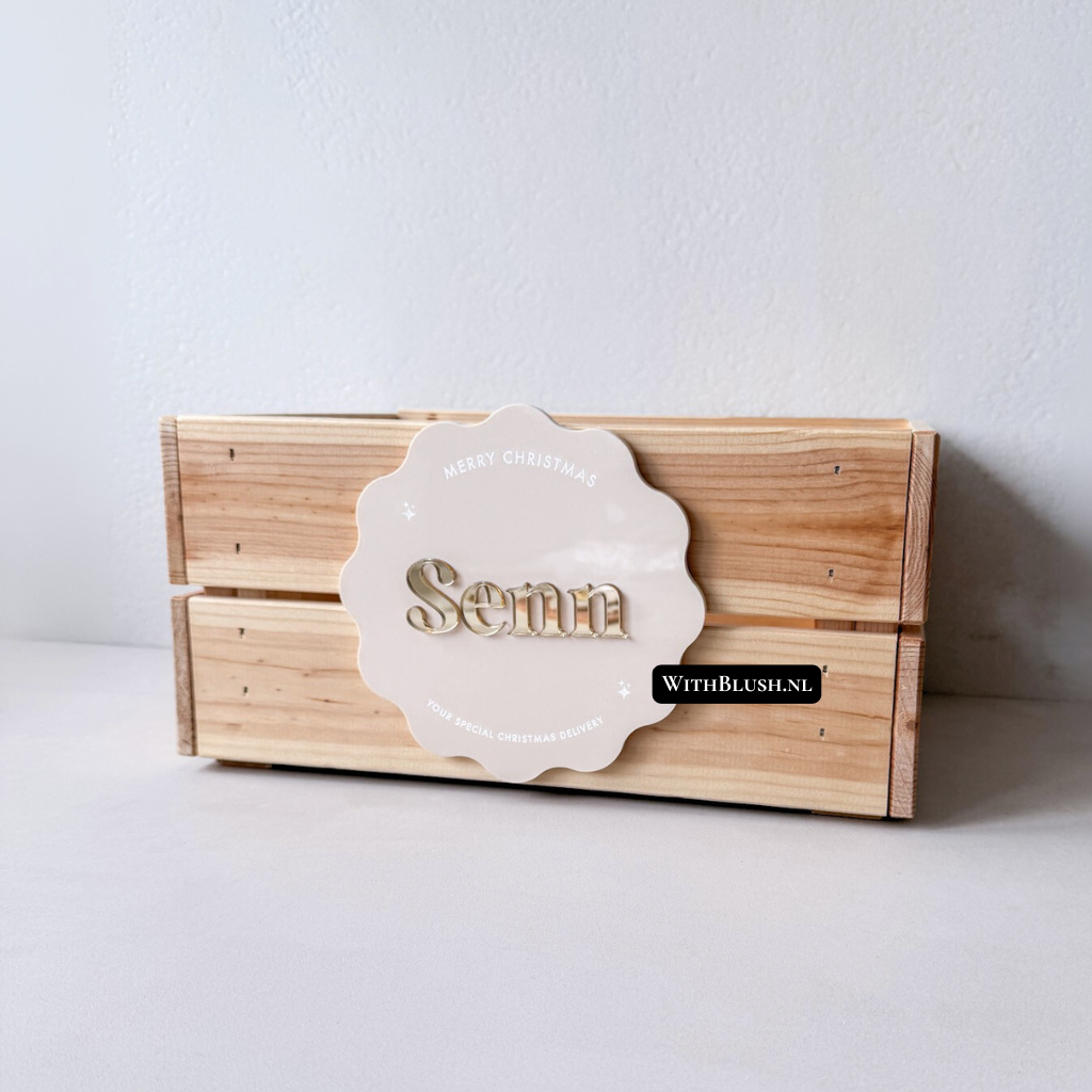 Gepersonaliseerde houten kerstkist met naamlabel, kan gevuld worden met kerstitems zoals een boek, pyjama en speelgoed. De kist heeft twee handvatten en staat onder een kerstboom als sfeervol cadeau.