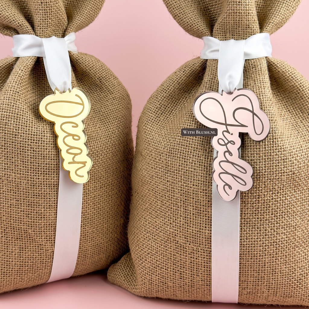 Twee gepersonaliseerde naamlabels. Aan het linkerzakje hangt een gouden gespiegelde acryl hanger met de naam "Ocean" in een sierlijk schrift. Aan het rechterzakje hangt een bronze hanger met de naam "Giselle".