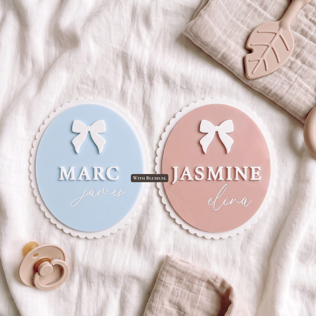 Twee unieke, gepersonaliseerde geboortebordjes voor baby's, liggend op een zachte doek. De namen 'MARC james' en 'JASMINE elina' zijn in wit aangebracht met een strikje. Perfecte decoratie voor de babykamer of als kraamcadeau.