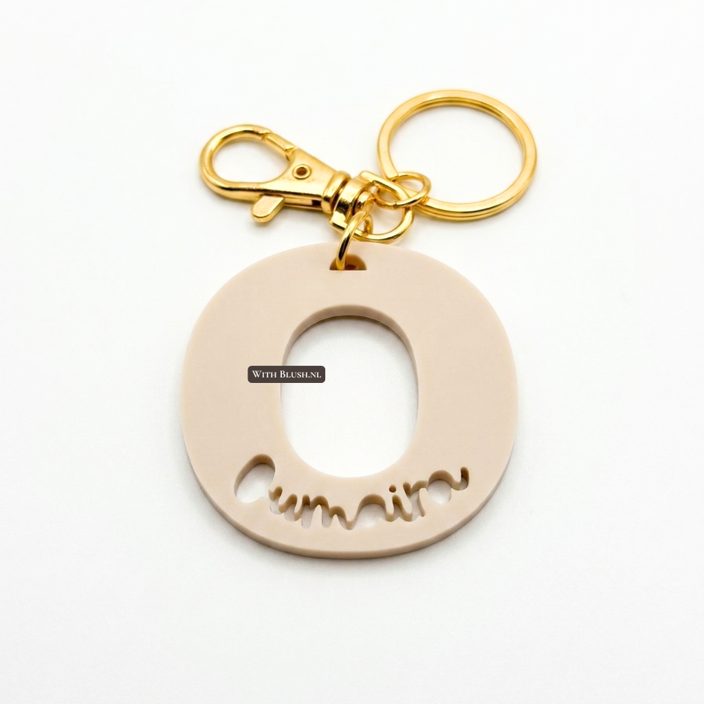 Een gepersonaliseerde sleutelhanger van mat beige plexiglas in de vorm van de letter O, met de naam 'Oumaira' eruit gesneden en een luxe gouden ring en clip.