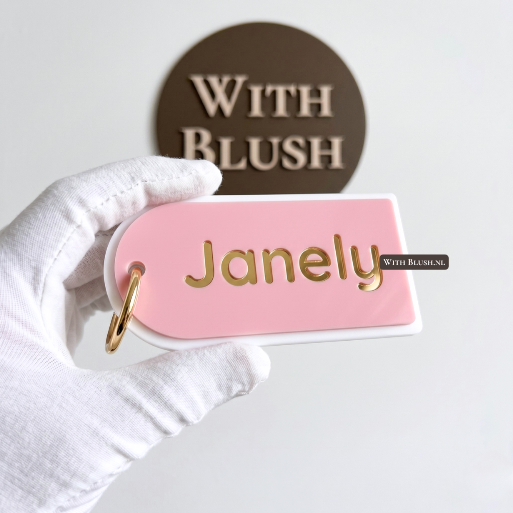 Gepersonaliseerde 3D sleutelhanger met naam in de vorm van een boog. Lichtroze acryl basislaag met de naam Janely in spiegelgoud, inclusief luxe gouden hardware. Gepresenteerd voor het With Blush logo.
