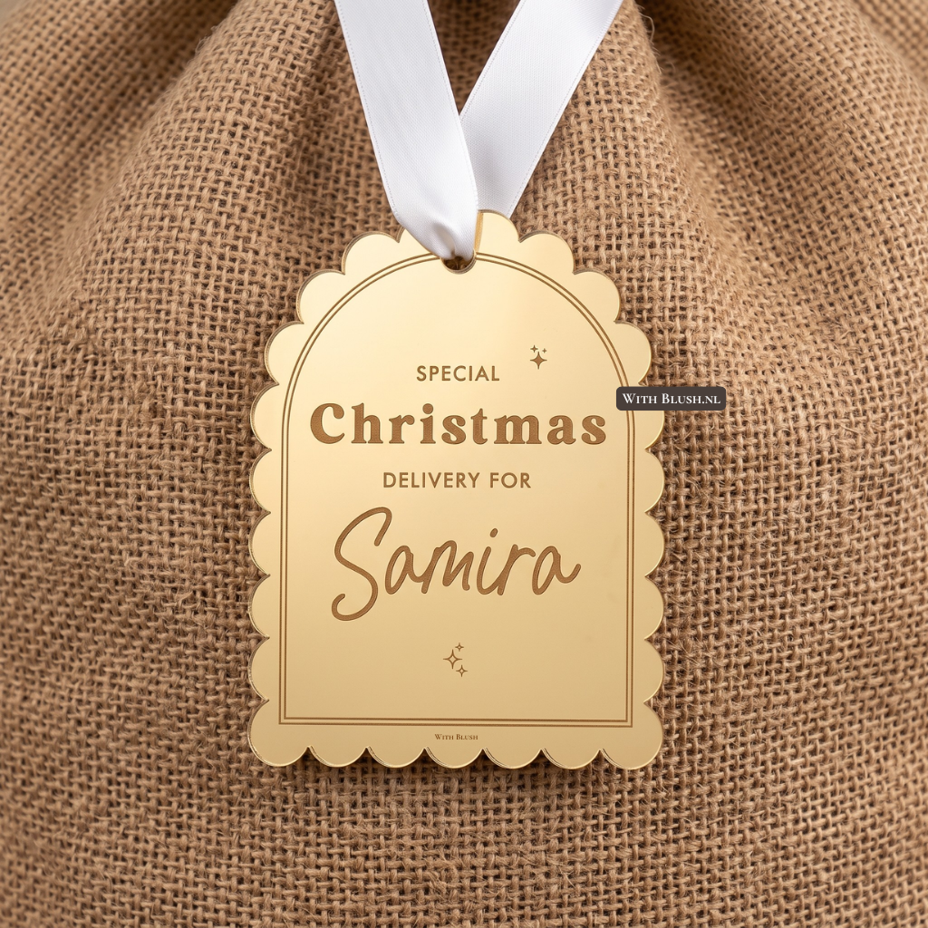 Een spiegelend goudkleurig geschulpt kerstcadeaulabel hangt aan een rustieke jutezak met een wit satijnen lint. Op het label staat de gegraveerde tekst "SPECIAL Christmas DELIVERY FOR Samira" met een klein sparkle-icoontje.