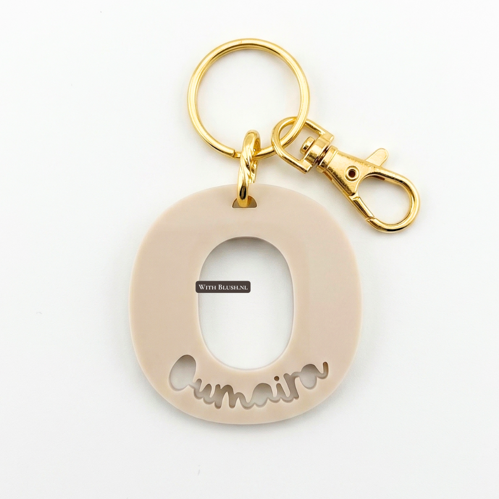 Close-up van een luxe mat beige acryl sleutelhanger initiaal O met uitgesneden naam en goudkleurige hardware, perfect als gepersonaliseerd cadeau.
