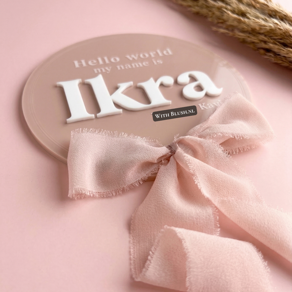 Gepersonaliseerd rond geboortebord van oudroze acryl voor baby Ikra met witte 3D letters en een roze lint. Een uniek naambordje voor de babykamer of als luxe kraamcadeau