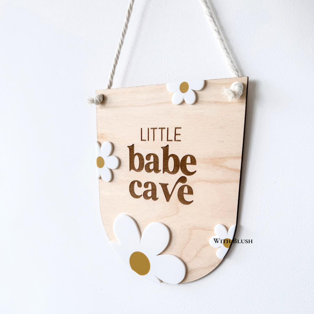 Houten of plexiglas Little Babe Cave vaantje met gegraveerde tekst en witte madeliefjes, perfect als wanddecoratie voor een babykamer of meisjeskamer.